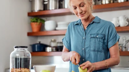 Older woman in a blue shirt slicing fruit, including apples and strawberries, on a cutting board in a modern kitchen. A jar of granola and a bowl are on the counter nearby.