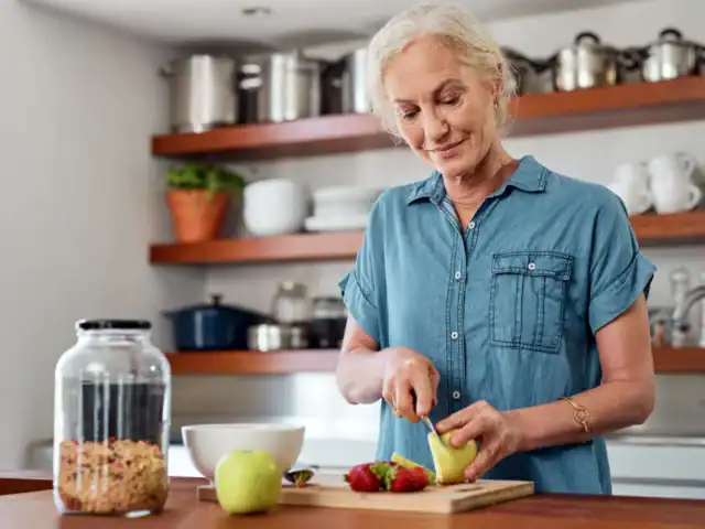 Older woman in a blue shirt slicing fruit, including apples and strawberries, on a cutting board in a modern kitchen. A jar of granola and a bowl are on the counter nearby.