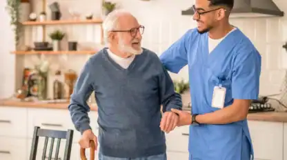 A young male nurse in blue scrubs assists an elderly man with a cane as they walk together in a bright kitchen.