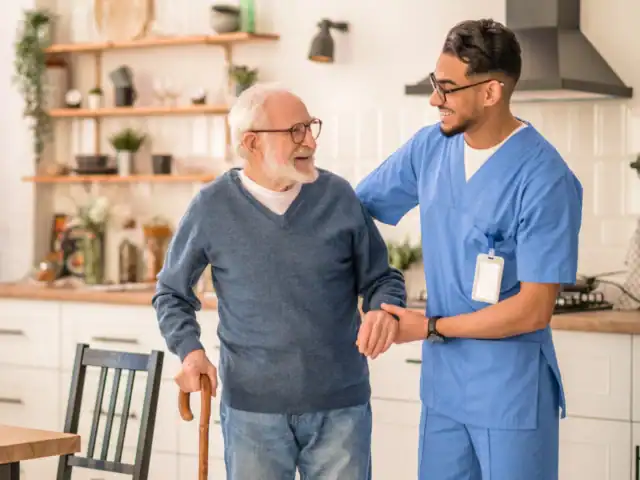 A young male nurse in blue scrubs assists an elderly man with a cane as they walk together in a bright kitchen.