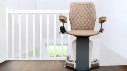 A beige stairlift chair on a rail is positioned at the top of a staircase next to a white railing, on a wooden floor.