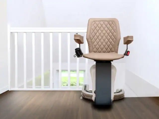 A beige stairlift chair on a rail is positioned at the top of a staircase next to a white railing, on a wooden floor.