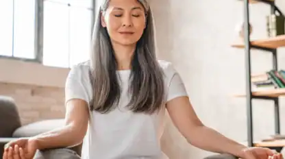 A woman with long gray hair sits cross-legged indoors, eyes closed, meditating with hands resting on her knees.