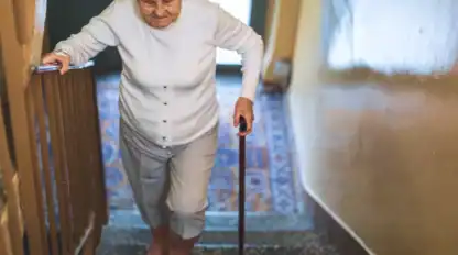 Elderly woman with white hair climbs stairs using a cane, holding the handrail, inside a building with patterned carpet and yellow walls.