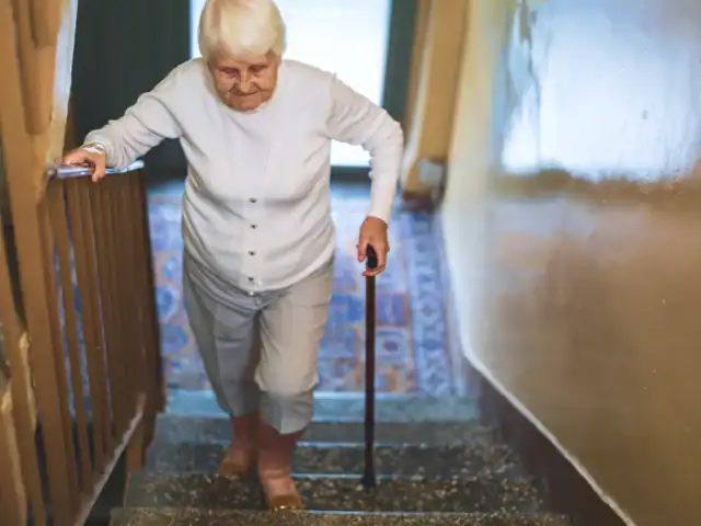Elderly woman with white hair climbs stairs using a cane, holding the handrail, inside a building with patterned carpet and yellow walls.