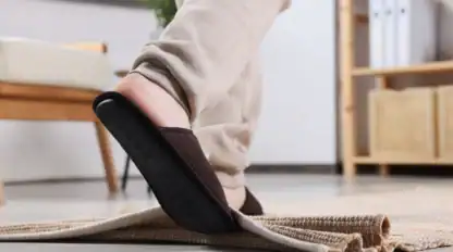 A person wearing slippers steps on a folded rug, causing it to lift from the floor in a home setting.