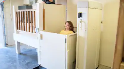 Two women use a residential wheelchair lift in a garage; one woman operates the lift while the other is seated inside it.