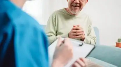An older man sits on a couch, smiling and clasping his hands, while a person in scrubs takes notes on a clipboard in the foreground.