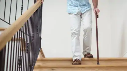 Man wearing white pants and blue plaid shirt walks down a wooden staircase using a cane, holding the railing with his other hand.