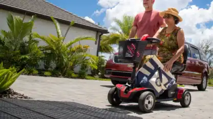 An older woman rides a mobility scooter with a tote bag while an older man walks beside her on a paved driveway near a red car and tropical plants.