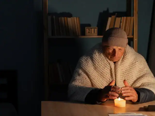 An older person in a knit hat and shawl sits at a table, warming their hands over a lit candle, with bookshelves in the background.