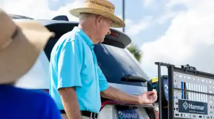 An older man in a straw hat and blue polo stands by a white SUV, operating a Harmar vehicle lift on a sunny day.