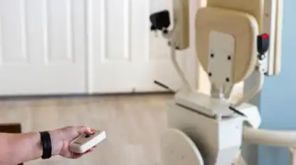 A person holds a remote control, operating a stair lift chair positioned at the base of a staircase in a home setting.