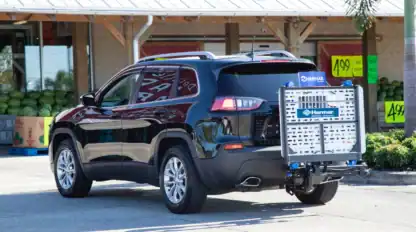 A black SUV with a silver Harmar lift attached to the rear is parked outside a store with watermelons and yellow sale signs visible.