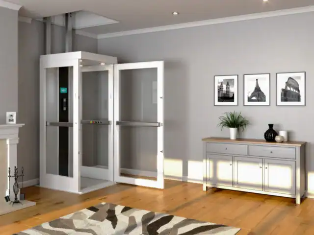 A home elevator with open doors is installed in a modern living room with wooden flooring, a rug, a sideboard, and framed photos on a gray wall.
