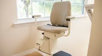 A beige stair lift chair is installed along a staircase next to a window with a view of palm trees outside.