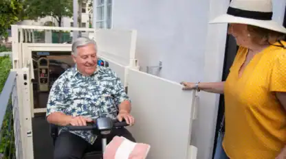An older man on a mobility scooter uses a vertical lift while a woman in a yellow top and sunhat holds the door open.