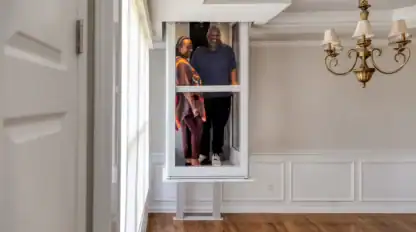Two people appear to be standing inside a large window frame elevated above the floor in an empty, sunlit room with hardwood floors and white walls.